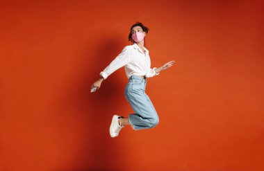 Kırmızı arka planda cep telefonuyla zıplayan maskeli neşeli genç bir kadın görüntüsü