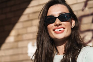 Dışarıda güneş gözlüğü takan, gülümseyen, iyimser genç bir kadının görüntüsü.