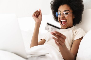 Yatakta kredi kartı ve dizüstü bilgisayarla online alışveriş yapan mutlu bir Afrikalı kadın.