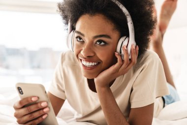 Çekici, genç Afrikalı bir kadın. Kulaklığı ve cep telefonuyla yatakta dinleniyor.