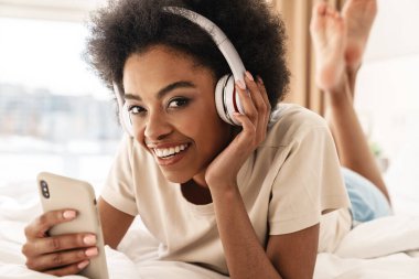 Çekici, genç Afrikalı bir kadın. Kulaklığı ve cep telefonuyla yatakta dinleniyor.