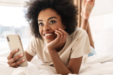 Genç gülümseyen Afrikalı kadın mesaj atıyor, yatak odasında yatıyor.