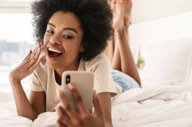 Mutlu genç Afrikalı kadın yatakta uzanırken selfie çekiyor.