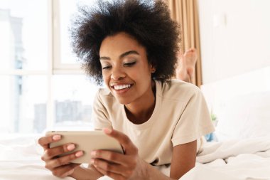 Mutlu genç Afrikalı kadın evde yatakta yatarken cep telefonuyla oyun oynuyor.