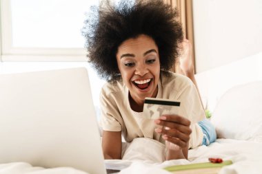 Yatakta kredi kartı ve dizüstü bilgisayarla online alışveriş yapan mutlu bir Afrikalı kadın.