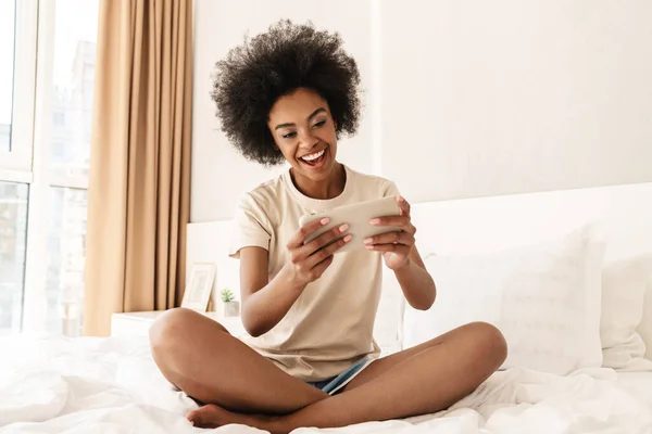 Счастливая Молодая Африканская Женщина Играет Игры Мобильном Телефоне Лежа Кровати — стоковое фото