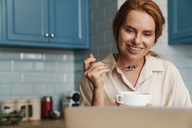 Neşeli kızıl kadının evde kahvaltı yaparken dizüstü bilgisayar kullanması.