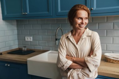 Mutfakta durup gülümseyen ve yan bakan mutlu kızıl saçlı kadın resmi.