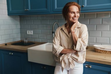 Mutfakta durup gülümseyen ve yan bakan mutlu kızıl saçlı kadın resmi.