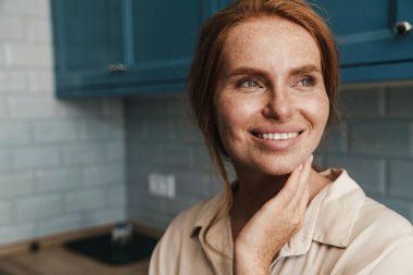 Mutfakta durup gülümseyen ve yan bakan mutlu kızıl saçlı kadın resmi.