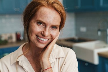 Mutfakta dikilirken gülümseyen ve kameraya bakan mutlu bir kızıl kadın resmi.
