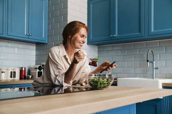 Imagem Mulher Ruiva Sorrindo Usando Celular Enquanto Come Salada Cozinha — Fotografia de Stock
