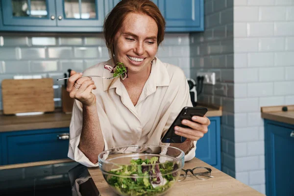 Imagem Mulher Ruiva Sorrindo Usando Celular Enquanto Come Salada Cozinha — Fotografia de Stock