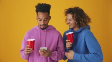 Odaklanmış bir adam, arkadaşı yanında dans ederken akıllı telefonunu kullanıyor. Stüdyonun turuncu arka planında izole bir şekilde.
