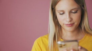 Pozitif sarışın bir kadının yakın planda çekilmiş görüntüsü stüdyodaki pembe arka planda tek başına duran akıllı telefonunu kullanıyor.