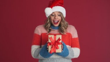 Noel Baba şapkalı genç, tatlı ve düşünceli bir kadın kırmızı duvar arkasında izole edilmiş hediye kutusunun içinde ne olduğunu tahmin etmeye çalışıyor.