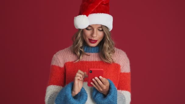 若いです楽観的な驚きの女性でクリスマスサンタ帽子使用して携帯電話隔離以上赤壁の背景 — ストック動画