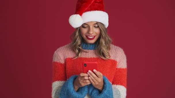 若いです陽気な幸せな女性でクリスマスサンタ帽子使用して携帯電話孤立以上赤壁の背景 — ストック動画