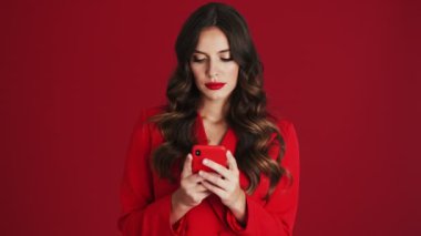 Kırmızı ceketli odaklanmış bir kadın, stüdyodaki kırmızı arka planda tek başına duran akıllı telefonunu kullanıyor.