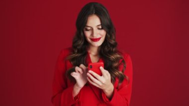 Kırmızı ceketli memnun bir kadın, stüdyoda kırmızı arka planda tek başına duran akıllı telefonuyla yazı yazıyor.