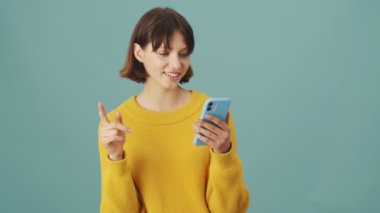 Mutlu bir kadın akıllı telefonuyla stüdyoda mavi bir arkaplanda tek başına daktilo yazıyor.