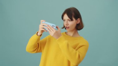 Neşeli bir kadın akıllı telefonuyla oyun oynuyor. Stüdyonun mavi arka planında tek başına duruyor.