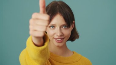 Kendine güvenen bir kadının yakın plan görüntüsü, stüdyonun mavi arka planında tek başına duran kameraya başparmak hareketi gösteriyor.