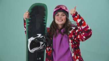 Mutlu bir genç kadın snowboardcu stüdyonun mavi arka planında tek başına dikilip baş parmak hareketi yapıyor.
