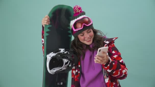 Позитивна Молода Жінка Сноубордист Використовує Свій Смартфон Стоячи Ізольовано Синьому — стокове відео