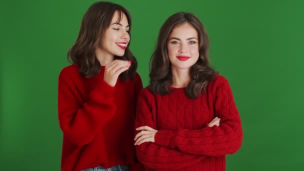 Две Улыбающиеся Девушки Красных Свитерах Делятся Секретами Стоящими Изолированными Зеленом — стоковое видео