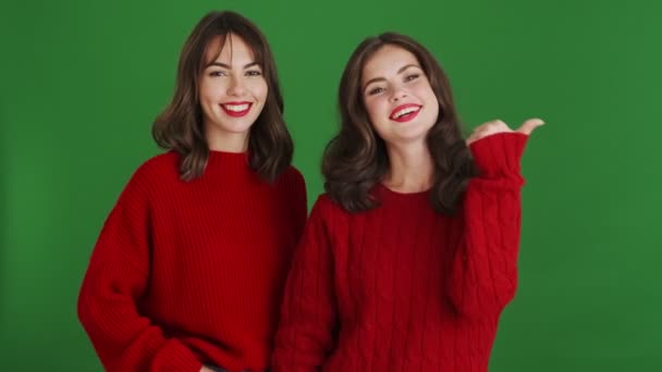Dwie Szczęśliwe Dziewczyny Czerwonych Swetrach Wskazują Stronę Stojącą Zielonym Tle — Wideo stockowe