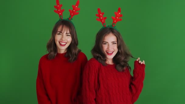 二つの美しいです女の子身に着けていますクリスマストナカイAntlersと赤セーターあります送信Aエアキスへザカメラスタンド絶縁上の緑の背景でザ スタジオ — ストック動画