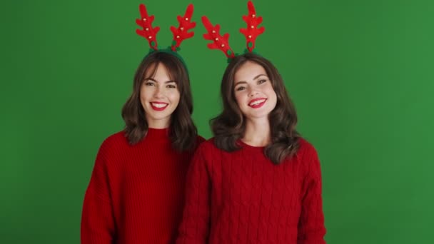 两名身穿圣诞驯鹿鹿角和红色毛衣的积极女孩站在摄影棚的绿色背景上 向摄像机打招呼 — 图库视频影像
