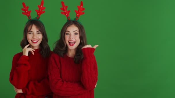 Two Positive Girls Wearing Christmas Reindeer Antlers Red Sweaters Pointing — Stock Video