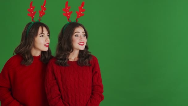 两个穿着圣诞驯鹿鹿角和红色毛衣的高兴女孩站在工作室的绿色背景上 看着旁边有可爱的东西 — 图库视频影像