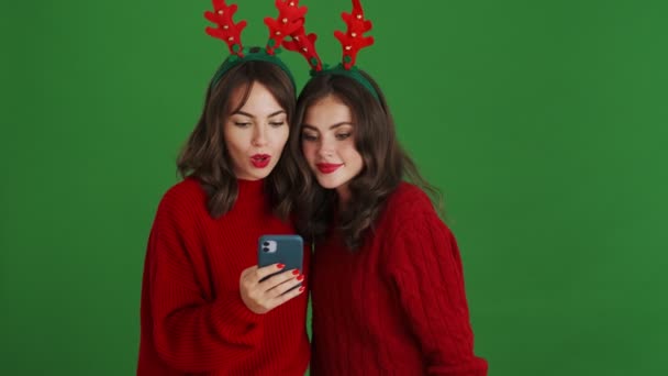 Дві Здивовані Дівчата Різдвяних Оленях Червоні Светри Використовують Смартфон Стоїть — стокове відео
