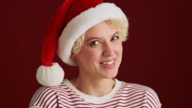 Mutlu genç kız Noel Baba şapkasıyla kameraya bakıyor. Kırmızı duvar arkasında izole edilmiş.