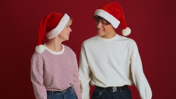 若いです楽観的な可愛いです女の子抱擁で彼女の兄でクリスマスサンタ帽子隔離されました以上赤壁の背景 — ストック動画
