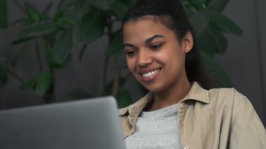 Gülümseyen Afrikalı Amerikalı bir kız kafedeki sandalyede oturan dizüstü bilgisayarını kullanıyor.