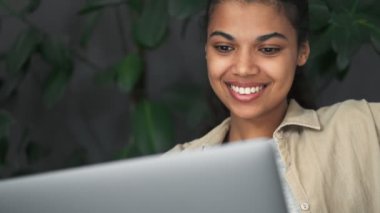 Memnun bir Afrikalı Amerikalı kız, kafenin içindeki sandalyede oturan dizüstü bilgisayarını kullanıyor.