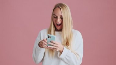 Stüdyoda pembe arka planda duran sarışın bir kadın akıllı telefonunu kullanıyor.