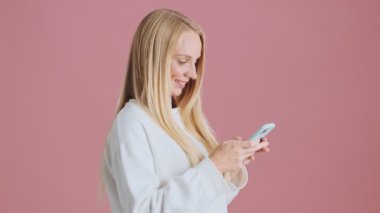 Mutlu bir sarışının yan görüntüsü akıllı telefonunu kullanırken stüdyonun pembe arka planında tek başına dikiliyor.