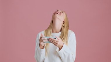 Gülen sarışın bir kadın akıllı telefonunu stüdyoda pembe arka planda yatay olarak dururken kullanıyor.