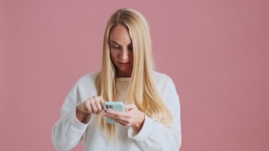 Konsantre olmuş sarışın bir kadın, stüdyoda pembe arka planda kapalı duran akıllı telefonunu kullanarak oyun oynuyor.