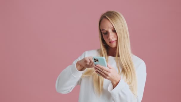 Glad Blond Kvinna Sveper Sin Smartphone Stående Isolerad Över Rosa — Stockvideo