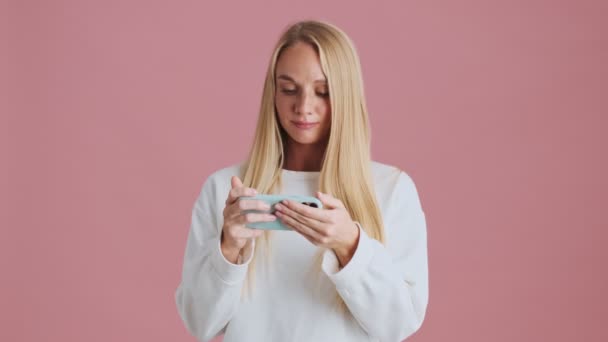一位专心致志的金发女人一边拿着智能手机 一边站在工作室的粉色背景上 与手机保持着横向隔离 — 图库视频影像