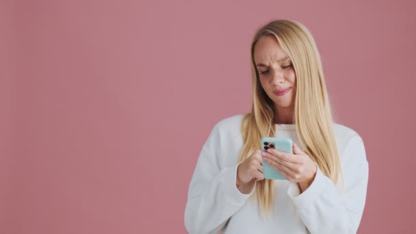 一位快乐的金发女人一边用智能手机 一边站在工作室里 孤零零地站在粉色的背景上 — 图库视频影像