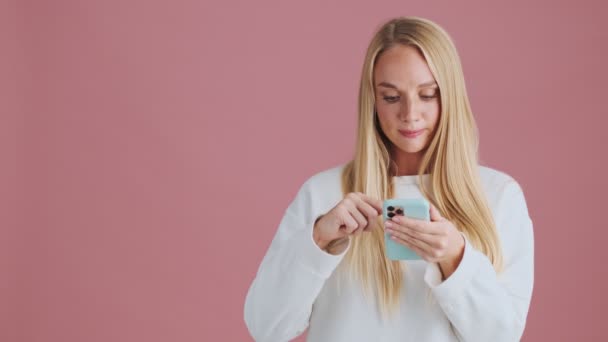 Задоволена Блондинка Вибирає Щось Використовуючи Свій Смартфон Стоїть Ізольовано Рожевому — стокове відео