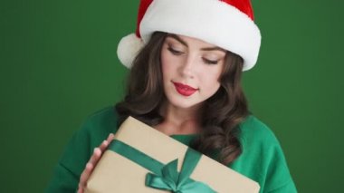Noel Baba şapkalı pozitif neşeli kız yeşil duvar arkasında izole edilmiş bir hediye tutuyor.
