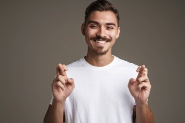 Şans dilemek için parmaklarını çaprazlayarak poz veren neşeli, tıraşsız bir adam görüntüsü. Gri arka planda izole edilmiş.